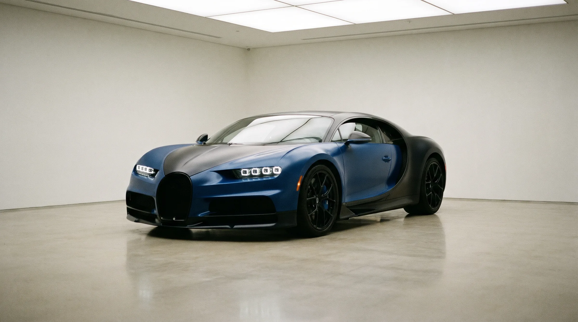 A blue and black hypercar parked in a white gallery garage under an overhead light panel